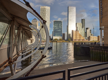 Canary Wharf hotel pier This urban architectural photograph shows the Canary Wharf hotel pier in London, United Kingdom, during a late afternoon in early autumn. The pier’s curved structure frames the scene, while the River Thames flows alongside it, reflecting light from the tall buildings of Canary Wharf in the background. The iconic skyscrapers, such as One Canada Square and other structures of the Canary Wharf financial district, are visible and create a striking cityscape. The photograph captures the urban atmosphere of London, highlighting the blend of contemporary architecture with the rippling water of the River Thames under autumn sunlight.